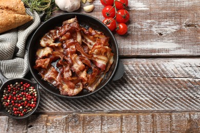 Delicious bacon slices in frying pan and products on wooden table, flat lay. Space for text