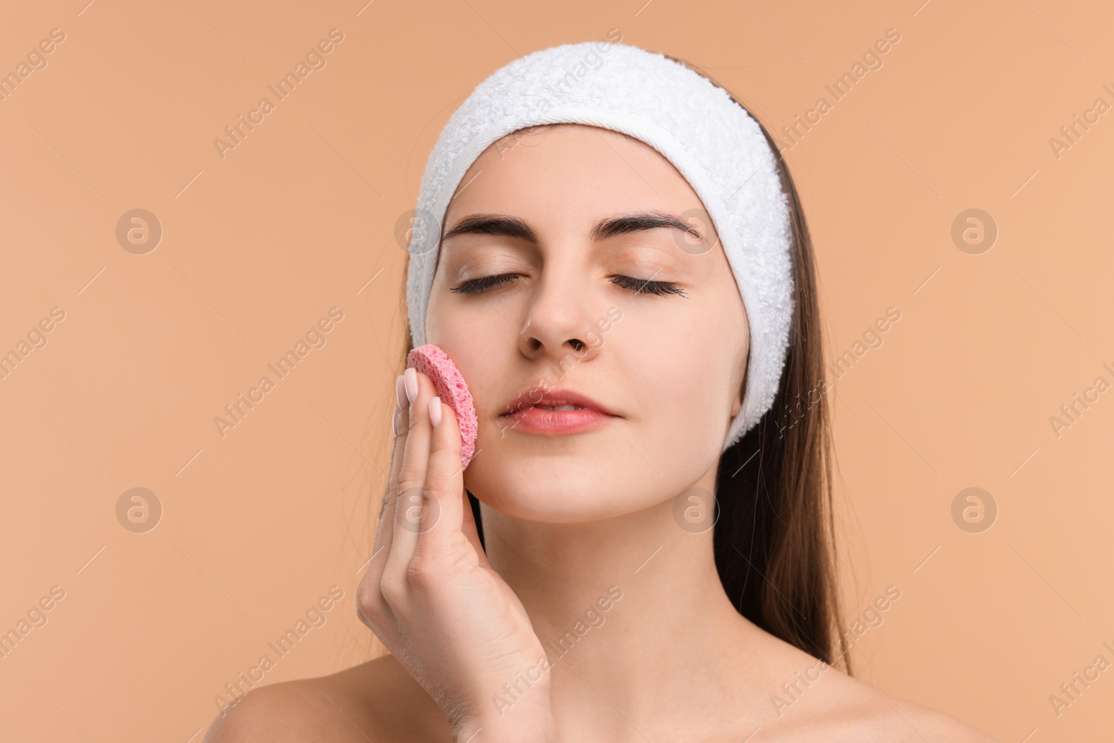 Photo of Young woman with headband washing her face using sponge on beige background