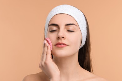 Young woman with headband washing her face using sponge on beige background