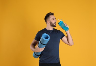 Handsome man with yoga mat drinking water on yellow background