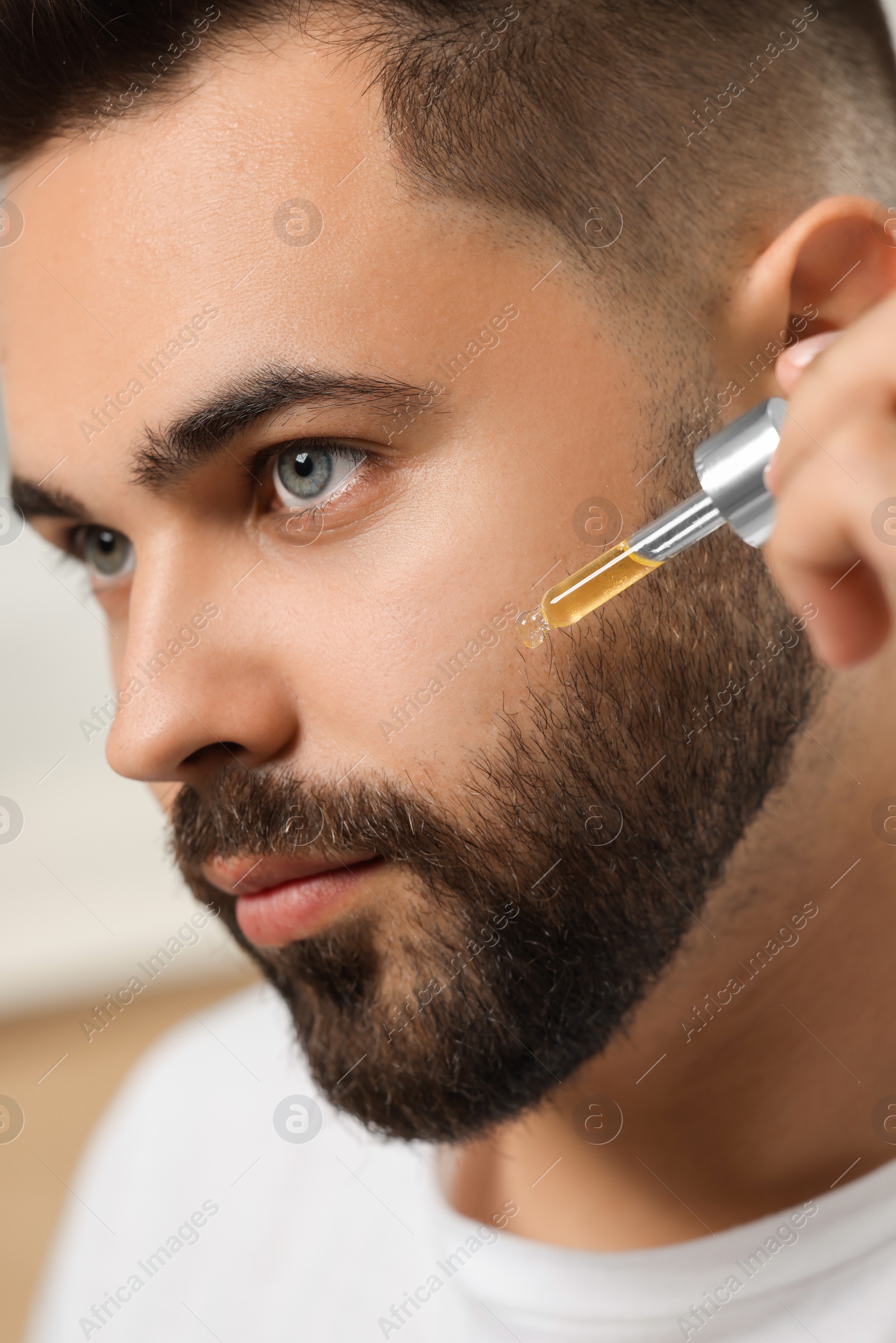 Photo of Handsome man applying cosmetic serum onto his face on blurred background, closeup