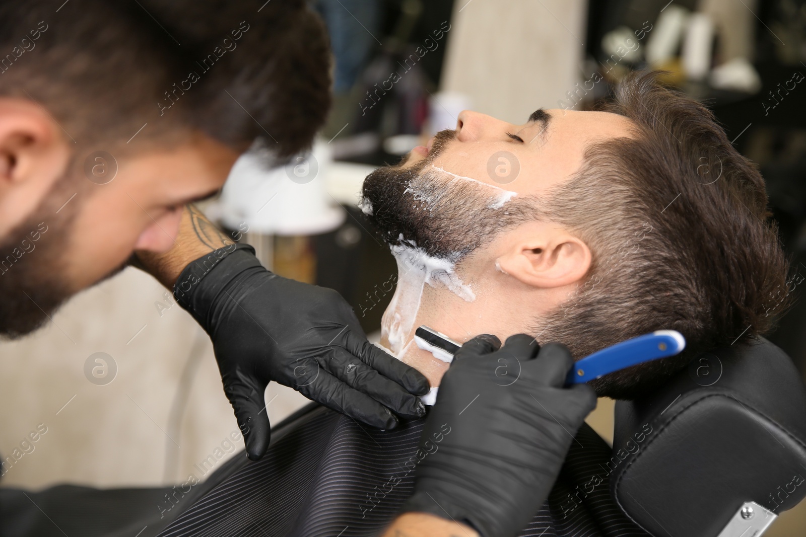 Photo of Professional hairdresser shaving client with straight razor in barbershop