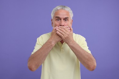 Embarrassed senior man covering mouth on purple background