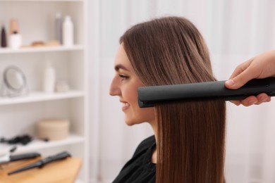 Photo of Hairdresser straightening woman's hair with flat iron in salon, selective focus