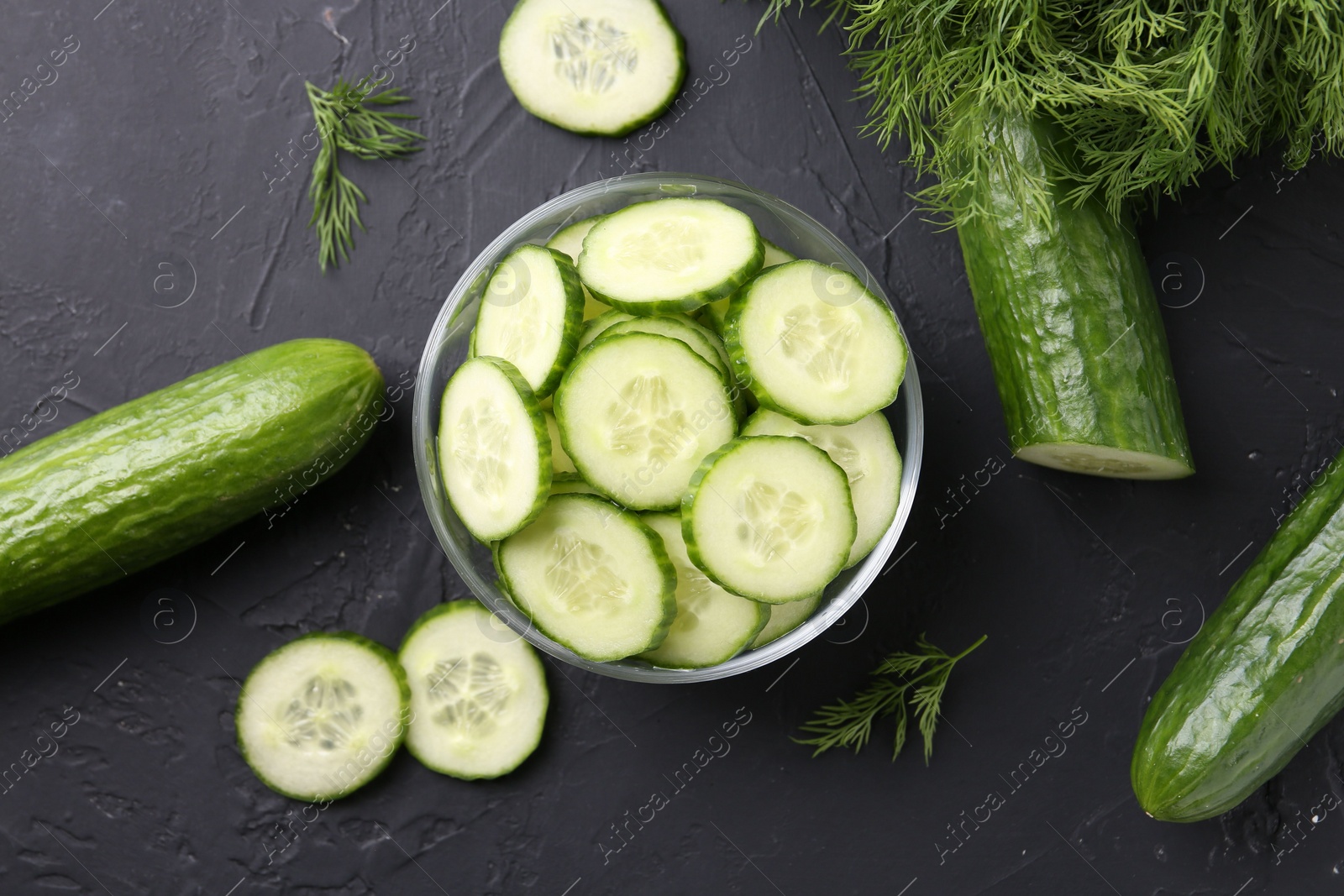 Photo of Cut cucumber in glass bowl, fresh vegetables and dill on dark gray textured table, flat lay