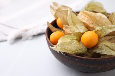 Photo of Ripe physalis fruits with calyxes in bowl on white table, closeup. Space for text