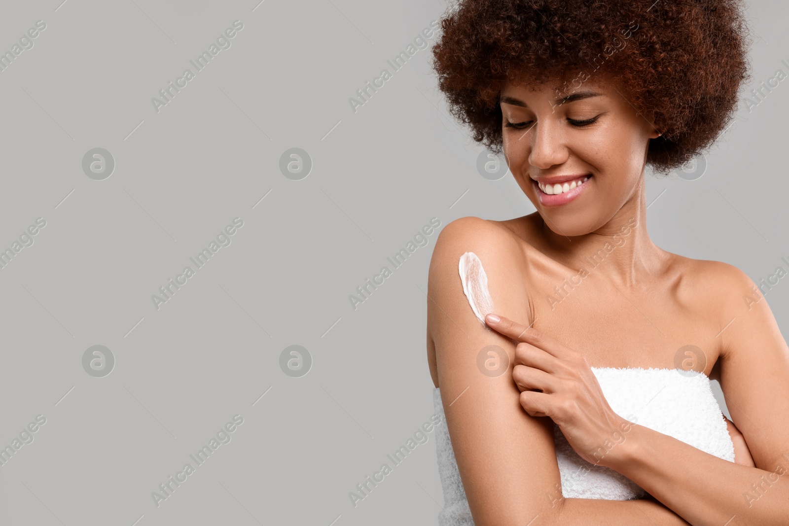 Photo of Beautiful young woman applying body cream onto arm on grey background, space for text