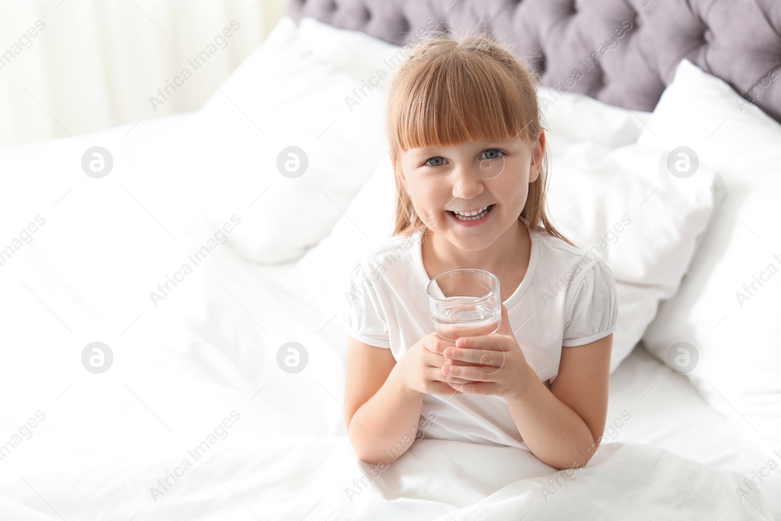 Photo of Cute little girl with glass of fresh water sitting in bed at home. Space for text