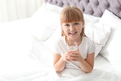 Photo of Cute little girl with glass of fresh water sitting in bed at home. Space for text