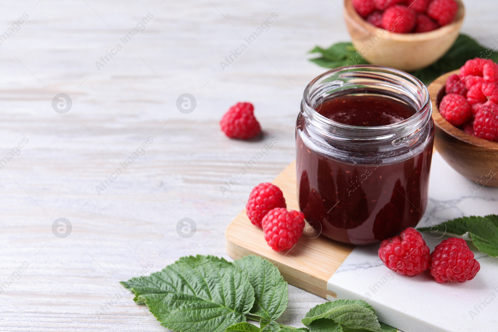 Photo of Delicious raspberry jam, fresh berries and green leaves on light wooden table. Space for text