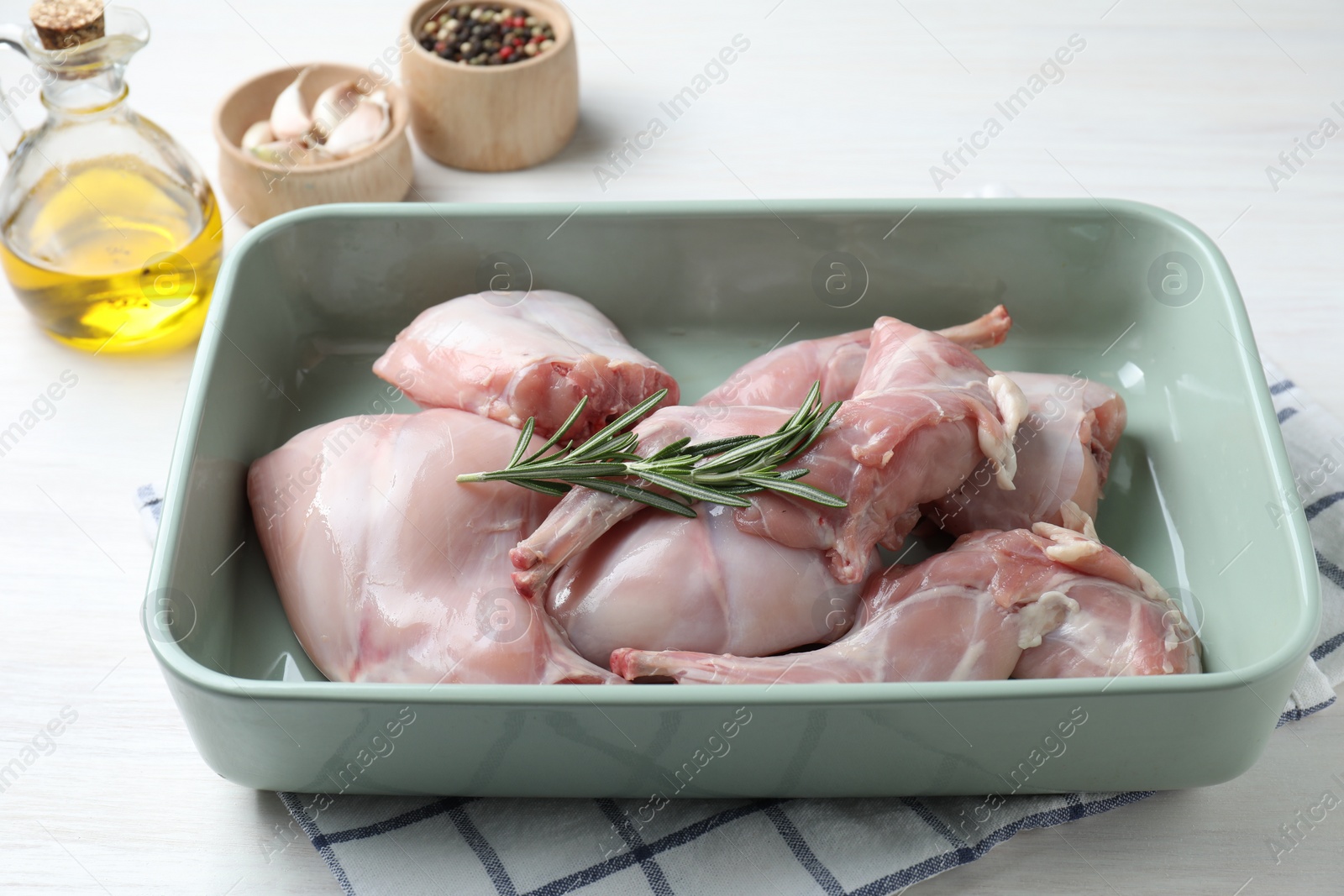 Photo of Raw rabbit meat and rosemary in baking dish on white wooden table, closeup