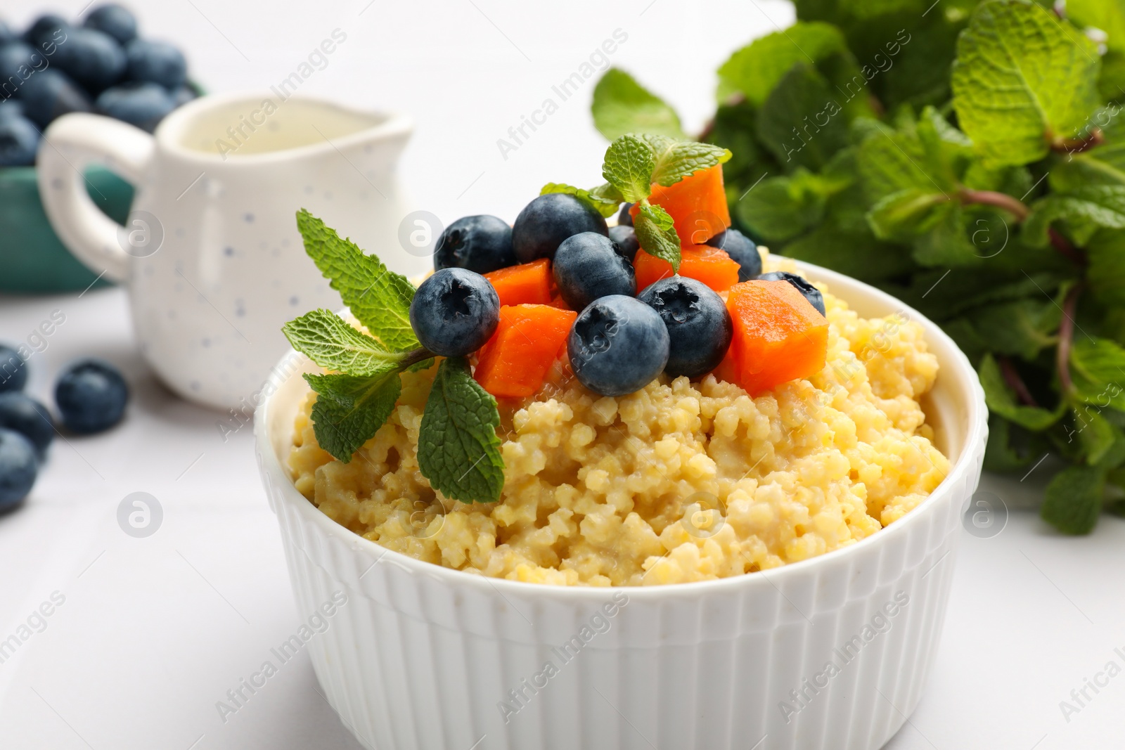 Photo of Tasty millet porridge with blueberries, pumpkin and mint in bowl on white tiled table, closeup