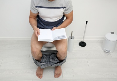 Young man with book sitting on toilet bowl in bathroom