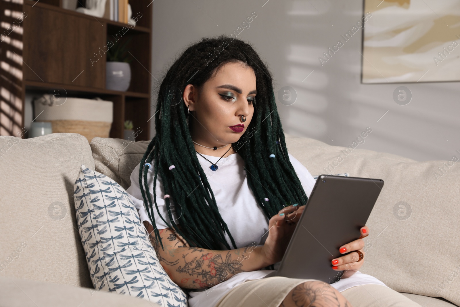 Photo of Beautiful young woman with tattoos on body, nose piercing and dreadlocks using tablet at home