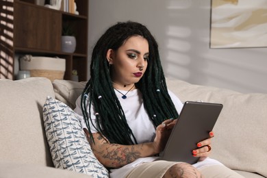 Beautiful young woman with tattoos on body, nose piercing and dreadlocks using tablet at home