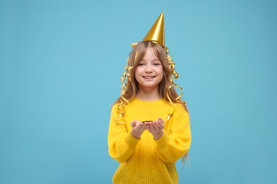 Happy little girl in party hat holding something on light blue background