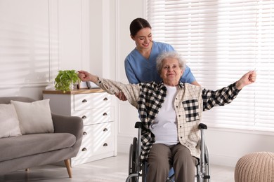 Photo of Senior woman in wheelchair doing physical exercise and young caregiver helping her indoors. Home health care service