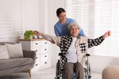 Senior woman in wheelchair doing physical exercise and young caregiver helping her indoors. Home health care service