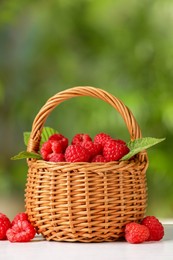 Photo of Wicker basket with tasty ripe raspberries and leaves on white table against blurred green background