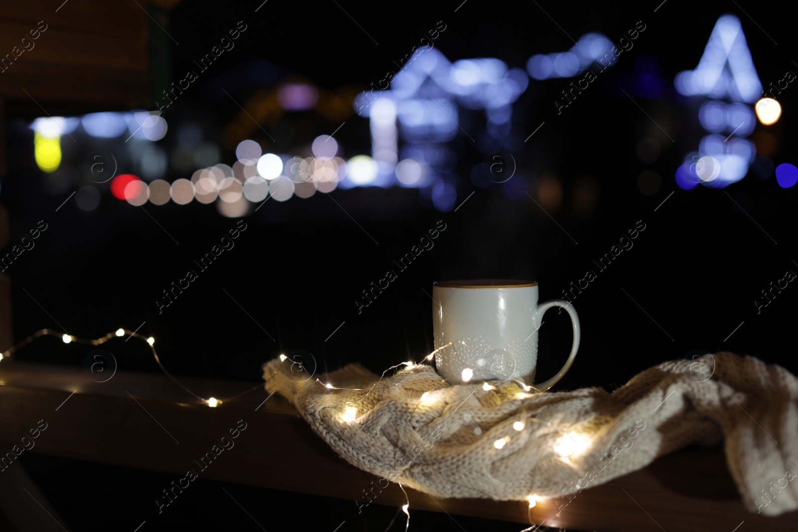 Photo of Cup of hot beverage, sweater and Christmas lights on wooden railing outdoors against blurred background, space for text. Winter night