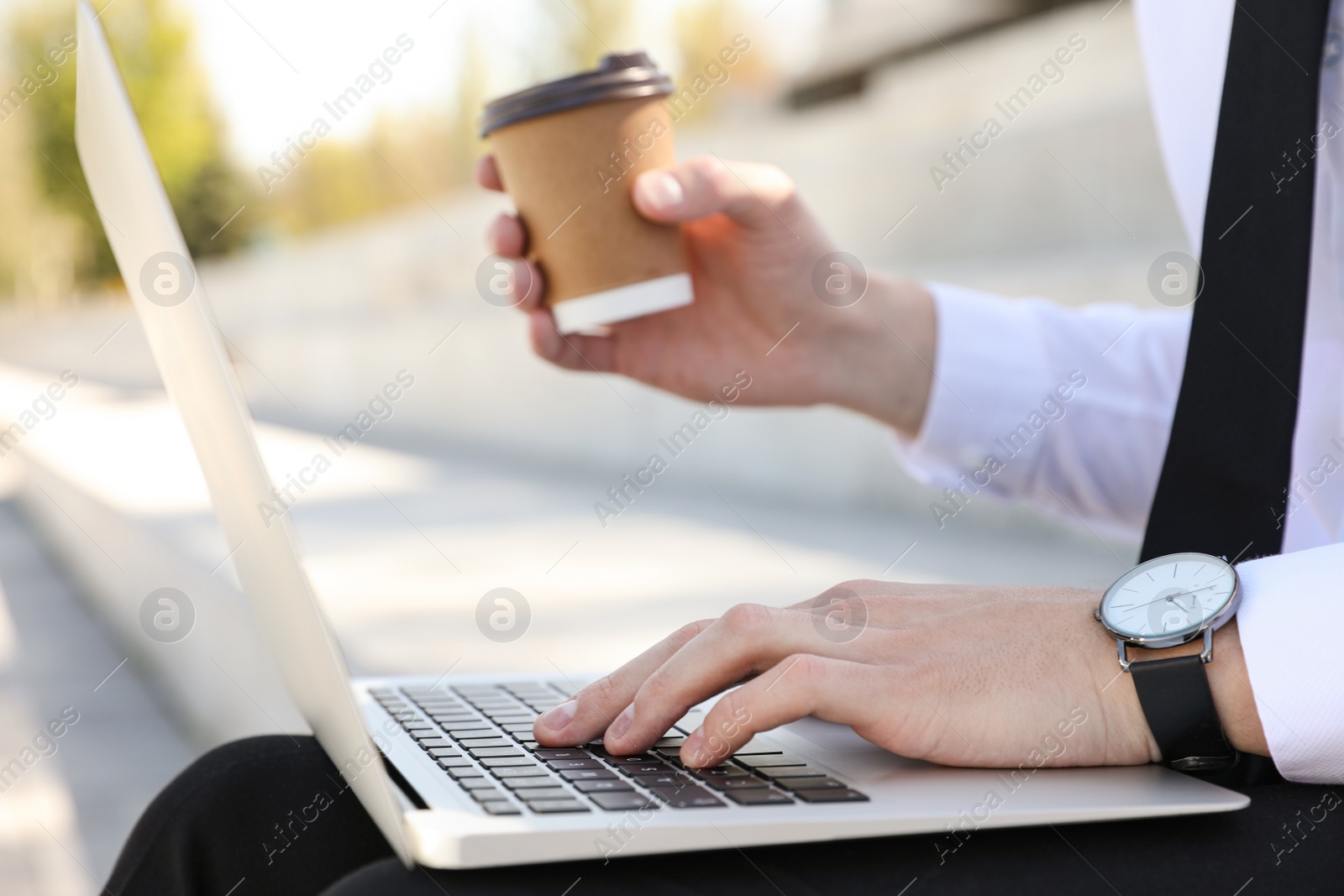 Image of Young man with paper cup of coffee working on laptop outdoors, closeup