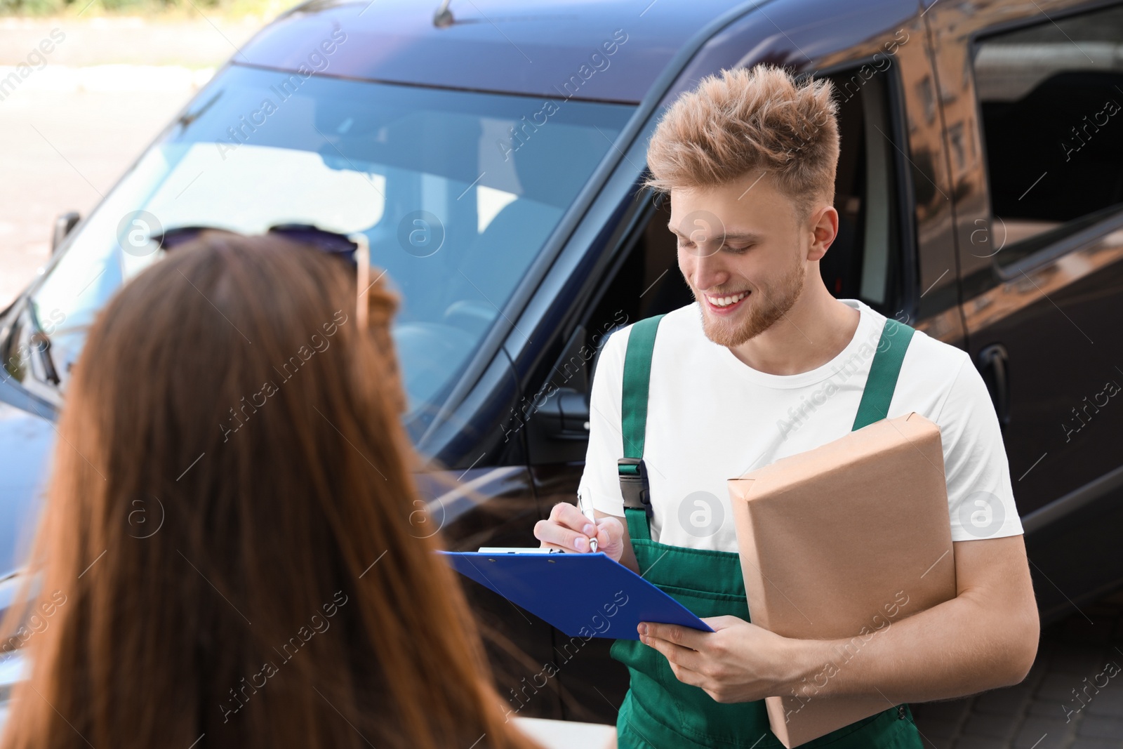 Photo of Young delivery courier with clipboard giving parcel to customer outdoors