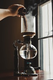Photo of Barista pouring coffee into vacuum maker at wooden table in cafe, closeup