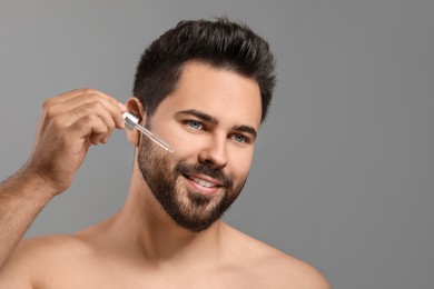 Handsome man applying cosmetic serum onto face on grey background, space for text