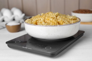 Electronic scales with uncooked pasta on white wooden table, closeup