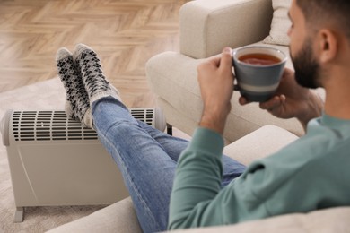 Photo of Young man with hot drink warming up near electric heater at home, focus on feet