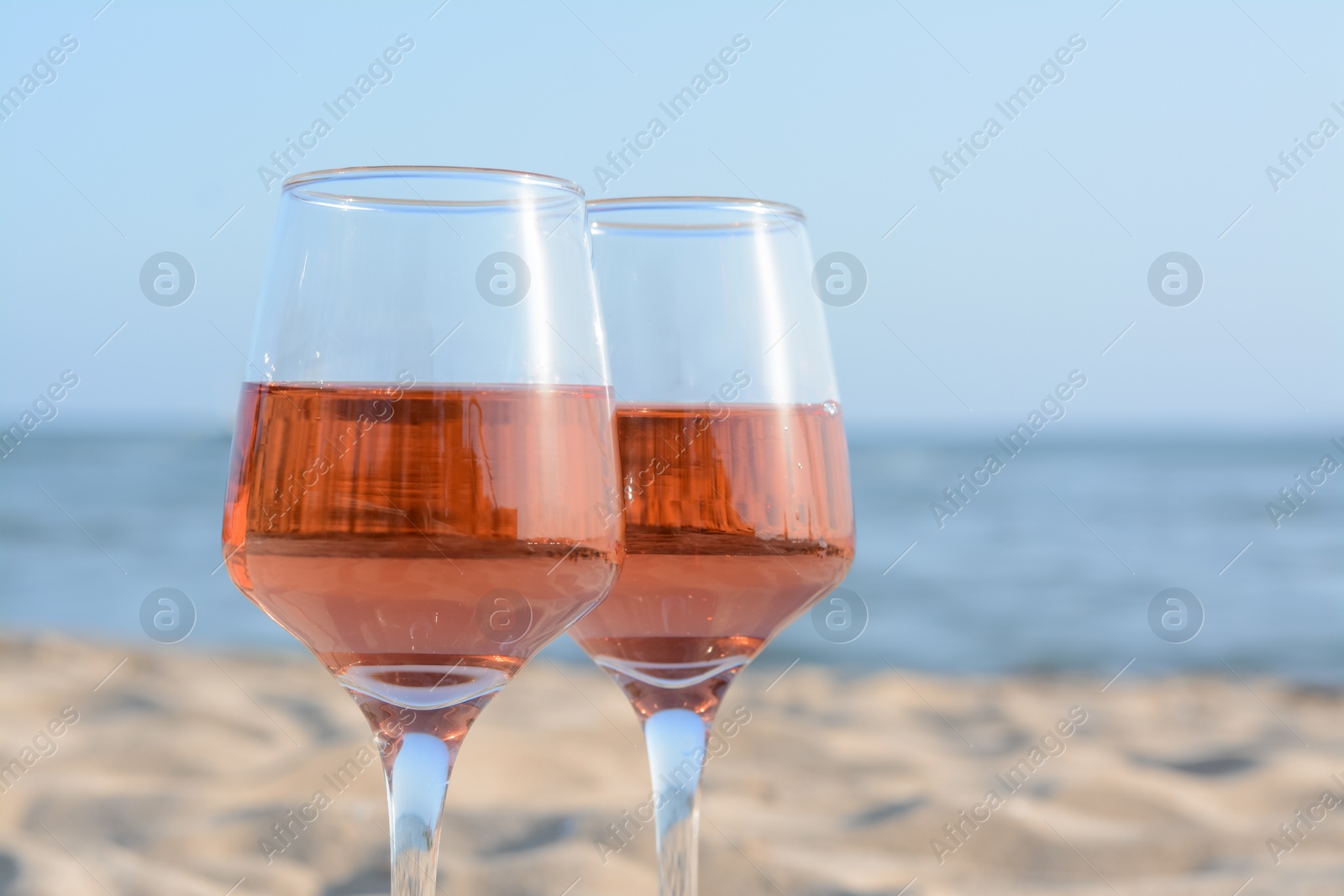 Photo of Glasses of tasty rose wine on sand near sea, closeup