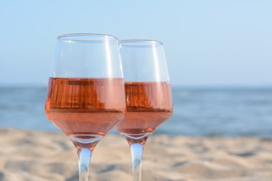 Glasses of tasty rose wine on sand near sea, closeup