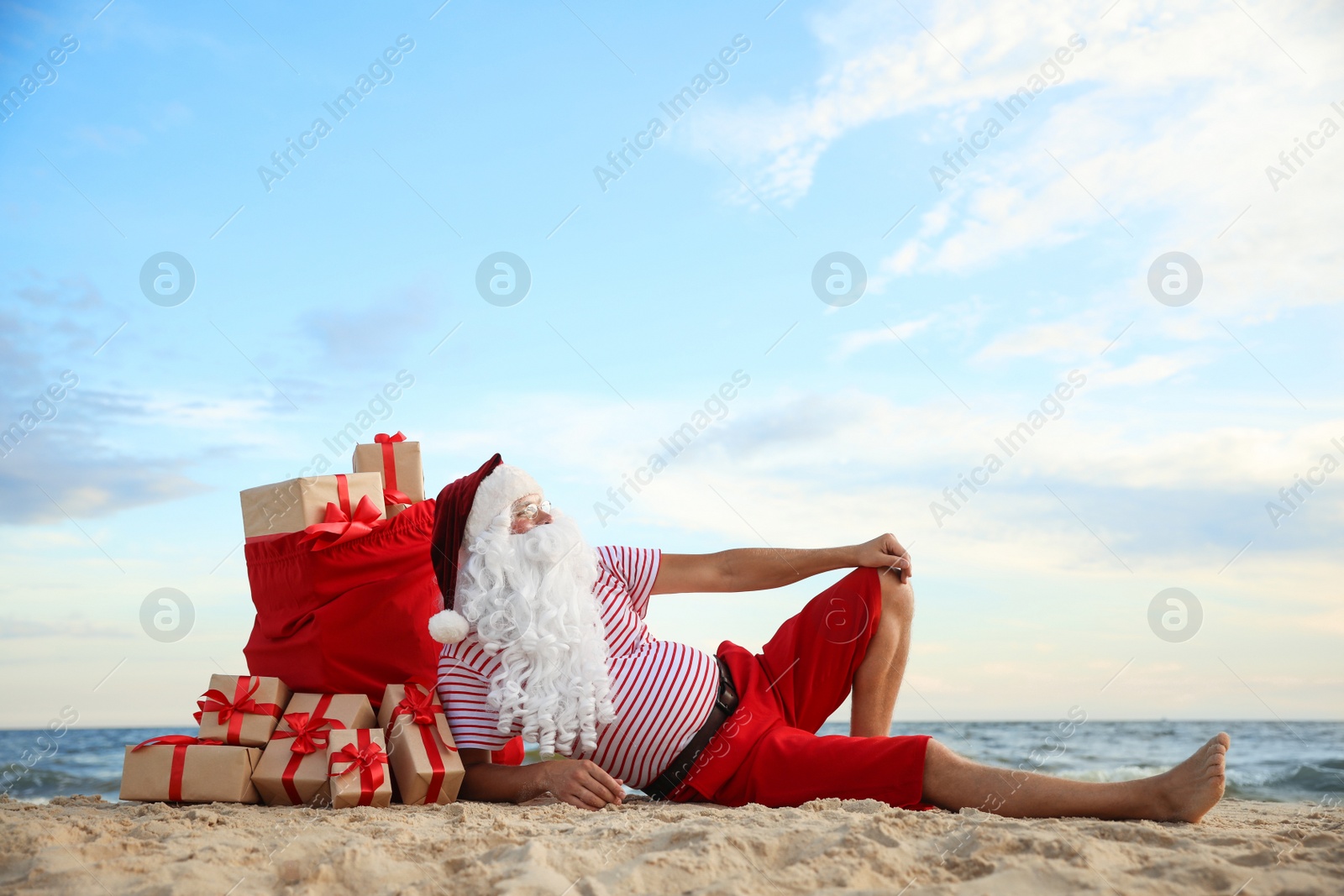 Photo of Santa Claus with bag of presents relaxing on beach, space for text. Christmas vacation