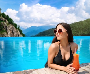 Image of Beautiful young woman with cocktail in outdoor swimming pool at luxury resort and beautiful view of mountains on sunny day