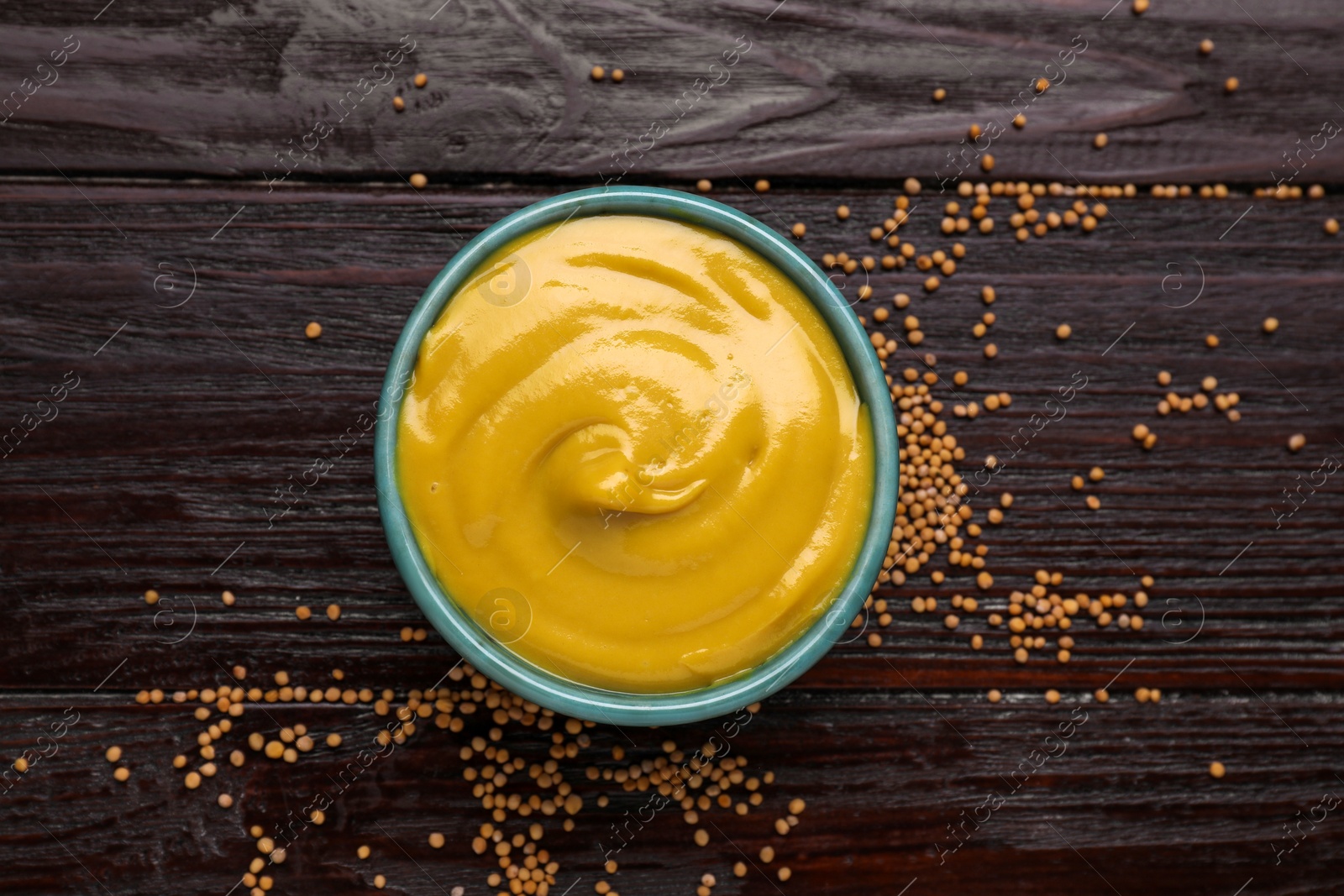 Photo of Sauce bowl with delicious mustard and seeds on dark wooden table, flat lay