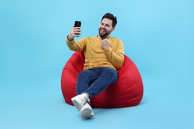 Happy young man using smartphone on bean bag chair against light blue background