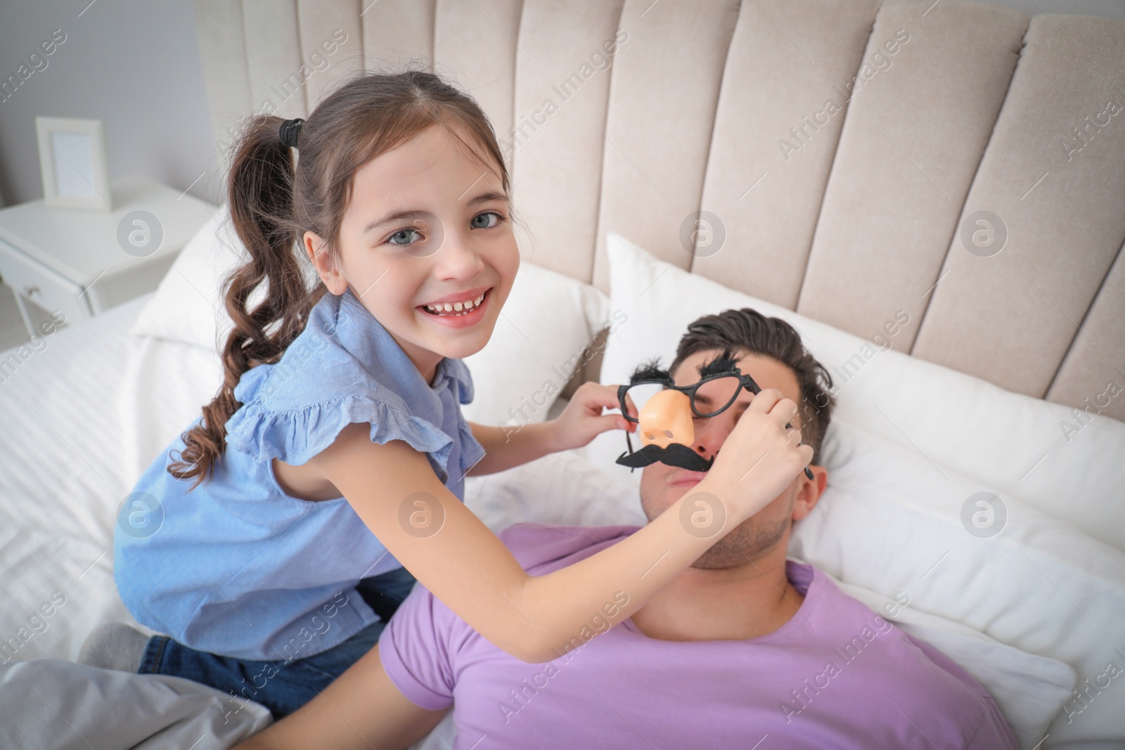 Photo of Cute little girl putting funny glasses on father while he sleeping in bed at home