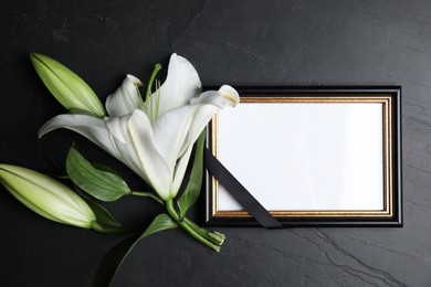 Photo of Funeral photo frame with ribbon and white lily on black table, flat lay. Space for design