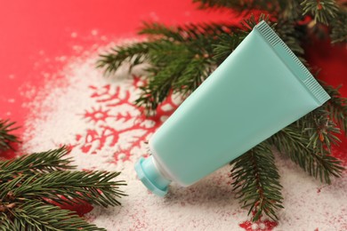Winter skin care. Hand cream near snowflake silhouettes made with artificial snow and fir branches on red background, closeup