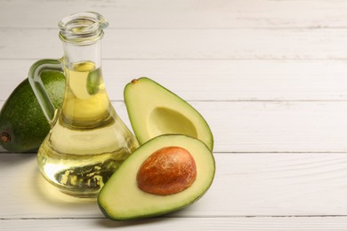 Glass jug of cooking oil and fresh avocados on white wooden table, space for text