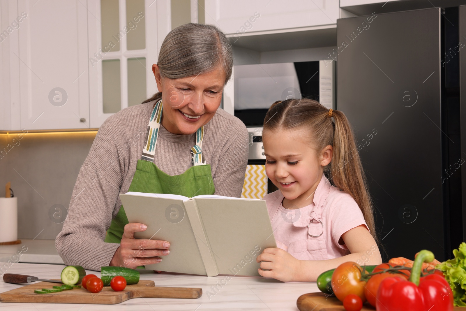 Photo of Cute little girl with her granny cooking by recipe book in kitchen