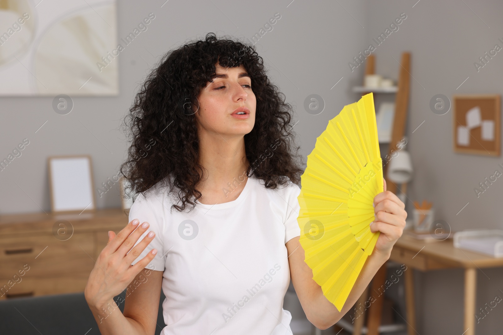Photo of Young woman waving yellow hand fan to cool herself at home