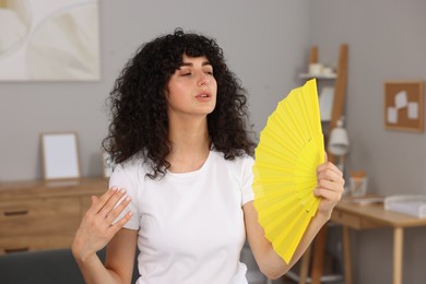 Young woman waving yellow hand fan to cool herself at home