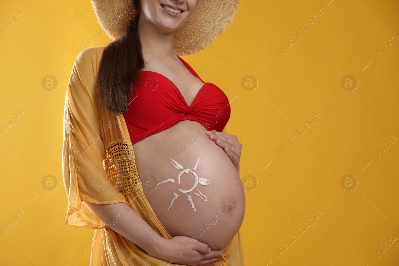 Photo of Young pregnant woman with sun protection cream on belly against yellow background, closeup. Space for text