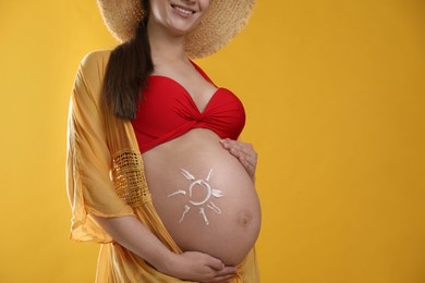Photo of Young pregnant woman with sun protection cream on belly against yellow background, closeup. Space for text