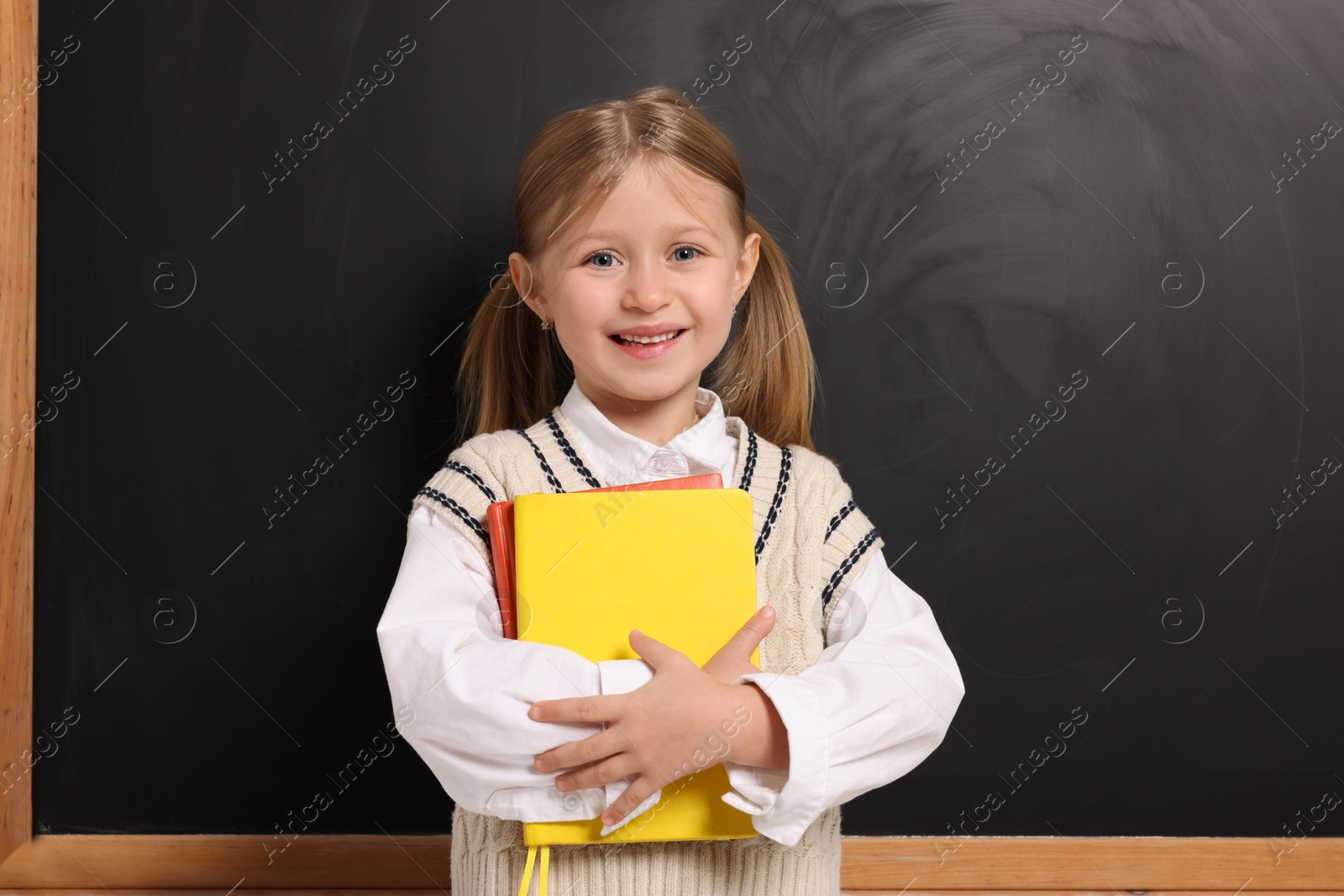 Photo of Happy schoolgirl with backpack and books near blackboard