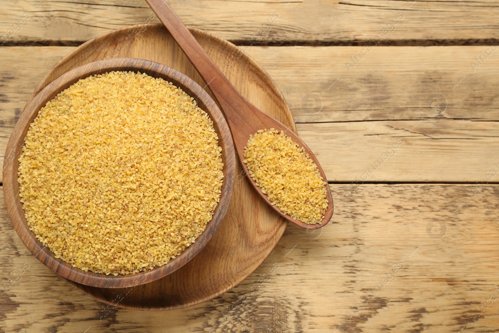 Photo of Bowl and spoon with uncooked bulgur on wooden table, top view. Space for text