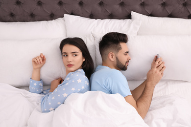Photo of Young man preferring smartphone over his girlfriend in bed at home, above view