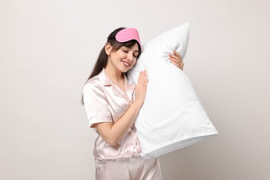 Happy woman in pyjama and sleep mask holding pillow on light grey background