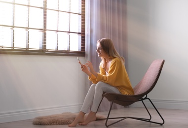 Photo of Young woman using smartphone near window with blinds at home. Space for text